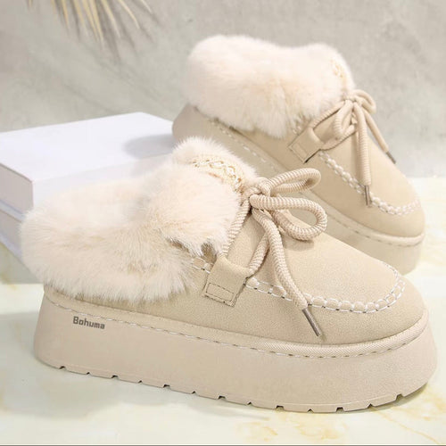 A pair of beige slip-on winter mule shoes with fluffy cream faux-fur trim, thick platform soles, and braided laces, shown on a light marble-like surface with a white box prop.