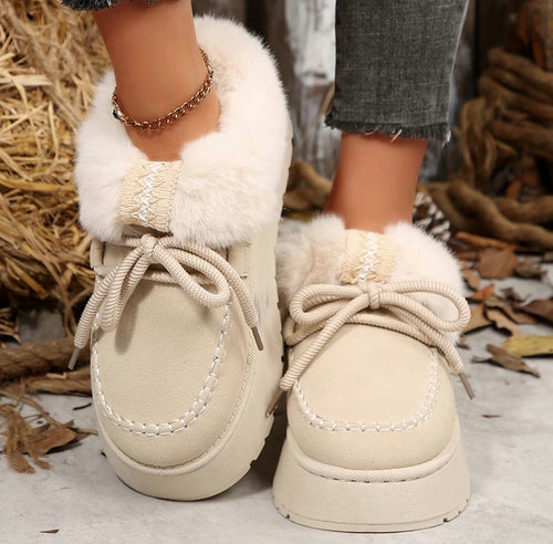 A pair of beige faux-suede winter boots with fluffy cream faux-fur trim and thick rope laces, shown on a person's feet outdoors on a stone surface with dry leaves and straw in the background.