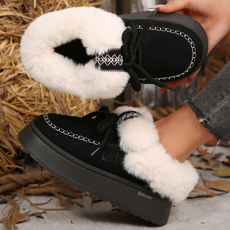 Black winter slipper boots with thick white faux fur lining and black laces, shown being worn with one held in hand against a rustic straw and leaf background.