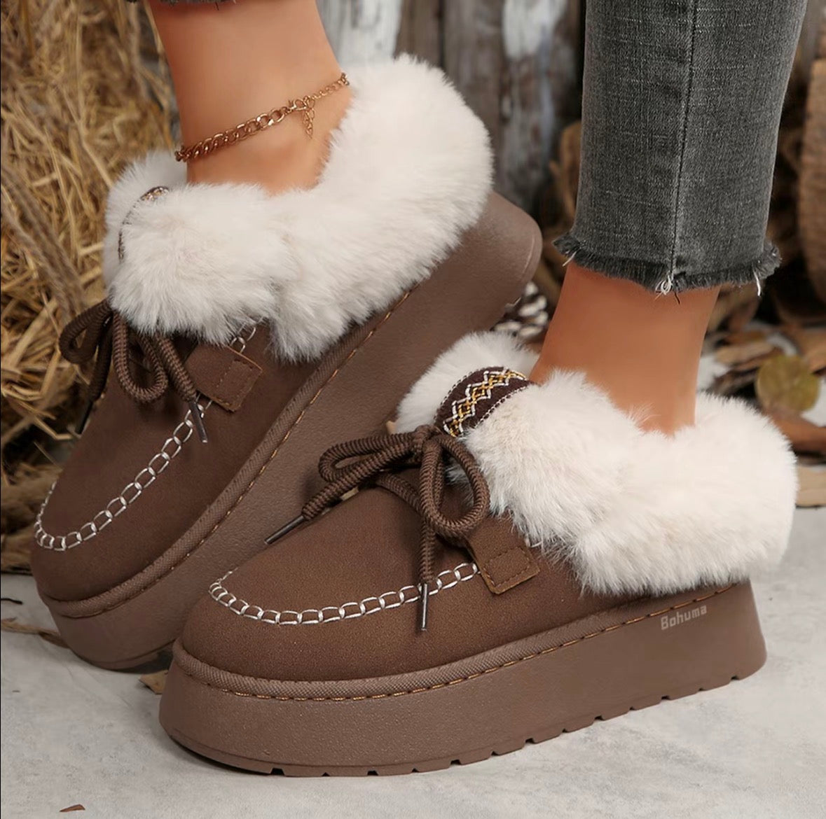 A pair of brown platform moccasin-style winter boots with thick white faux-fur trim, shown being worn on feet with gray jeans visible. The boots have brown laces, decorative stitching, and a chunky sole.